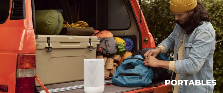 A man loads his backpack into the back of a van tailgate beside a Sonos Move 2 portable speaker in white, ready for a journey or outdoor activity.