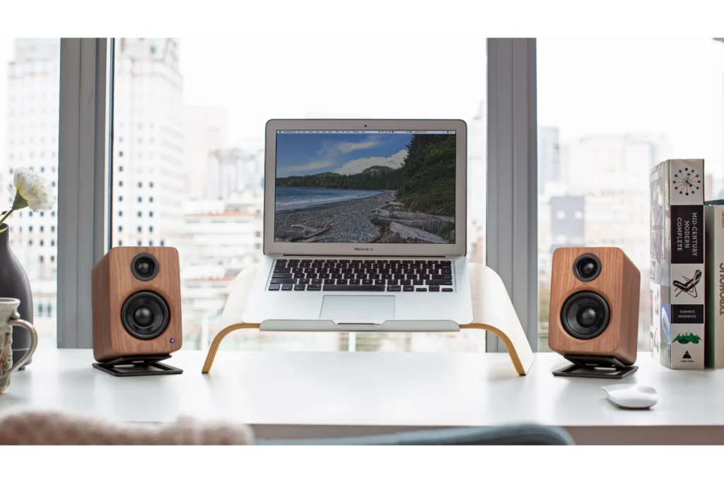 Kanto YU2 100W Speakers on a desk in the walnut colour way, clean desk beside matching laptop stand with view of city behind