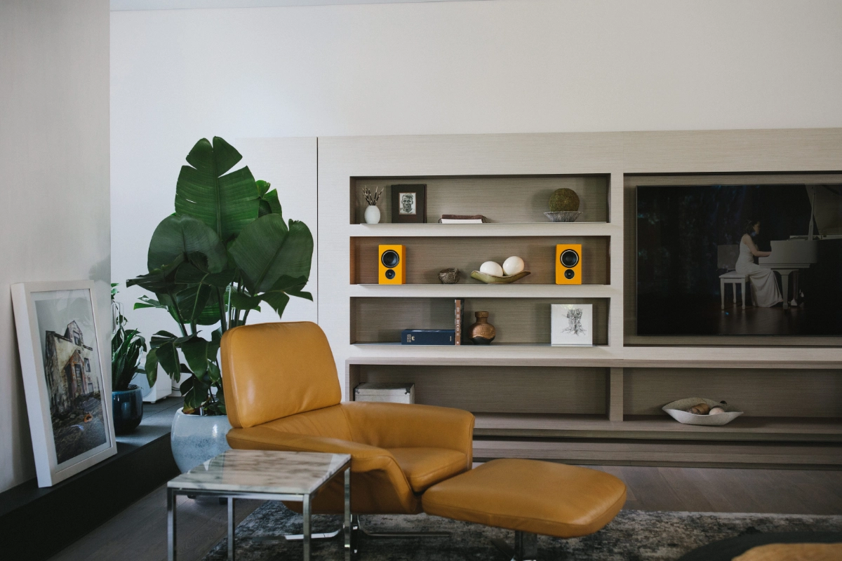 A stylish living room showcasing a yellow chair, a television, and yellow PSB Alpha iQ Speakers on a bookshelf.