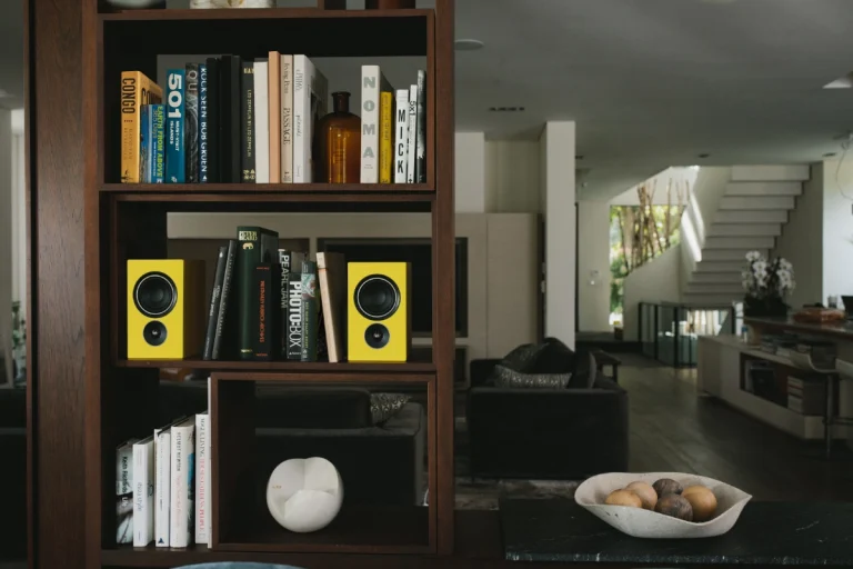 A pair of PSB Alpha iQ Speakers Pair in yellow flash boldly on a bookshelf in a dark living room.