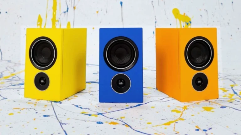 3 PSB Alpha Q bookshelf speakers in different colourways, yellow, blue and orange, with paint splatter in background.
