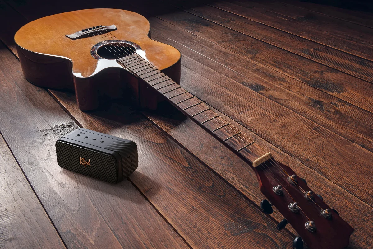 A guitar rests beside a Klipsch Nashville Portable speaker on a rustic wooden floor, creating a cozy atmosphere for music lovers.