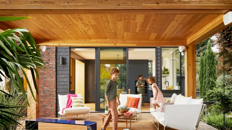 A family relaxes on a patio with a wooden ceiling, with Sonos by Sonance outdoor speakers mounted on the wall.