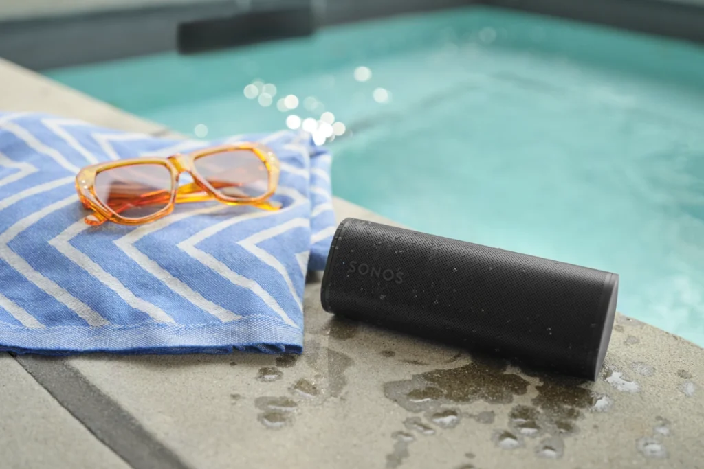 A black Sonos Roam 2 portable speaker, sunglasses, and a towel are placed beside a sparkling pool.