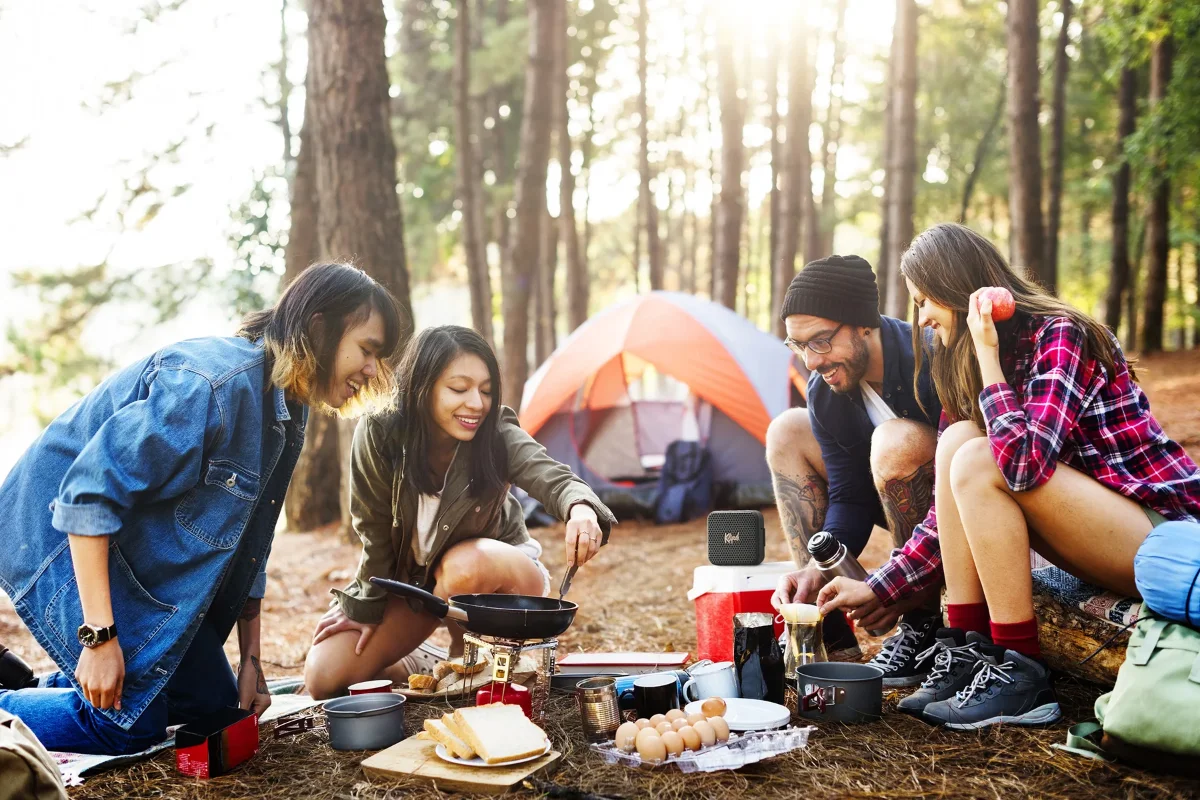 Four friends joyfully cooking a meal together in a serene forest setting, surrounded by trees and nature's beauty, enjoying music on the Klipsch Austin Portable speaker