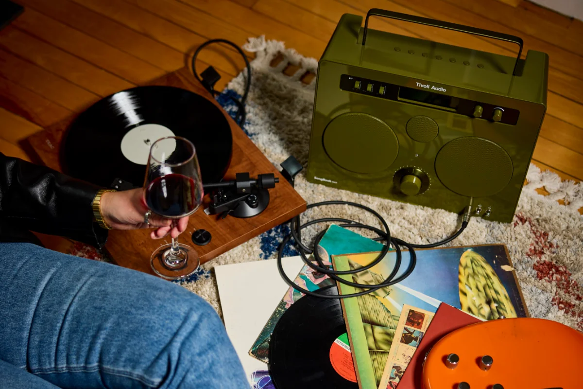 A person enjoys a glass of wine beside a Tivoli Songbook Max and record player, creating a cozy atmosphere.
