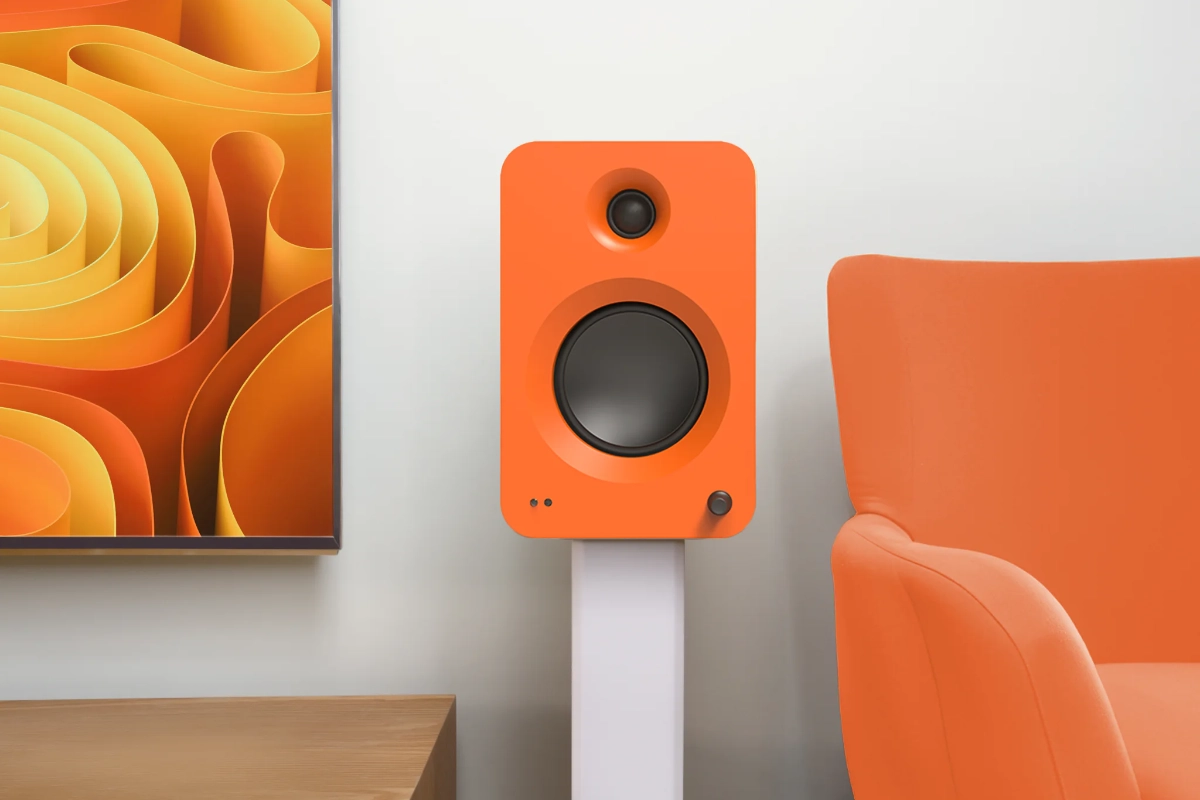Kanto REN speaker in matte orange, on white speaker stand beside orange chair and Orange TV