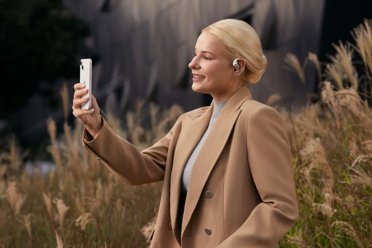 Woman wearing a Shokz OpenFit Pro earbud while taking a photo on her phone outdoors.