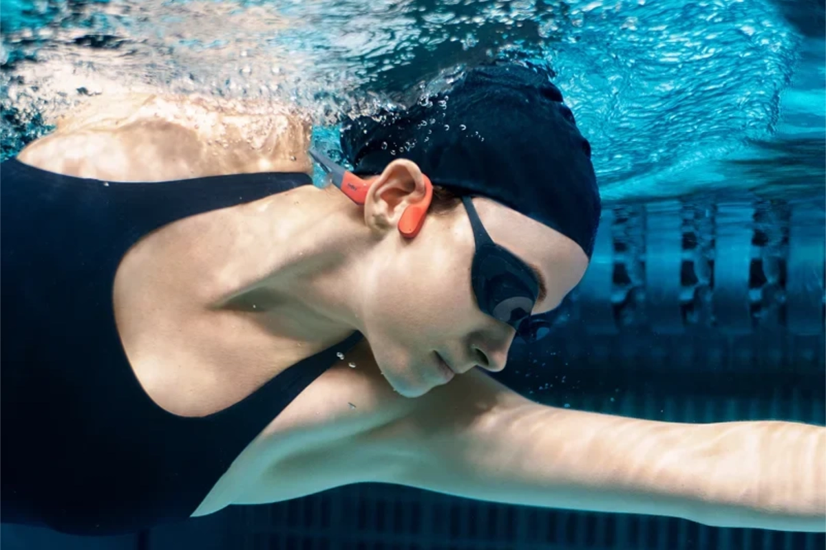 Woman swimming underwater in a pool while wearing orange Shokz OpenSwim Pro headphones.